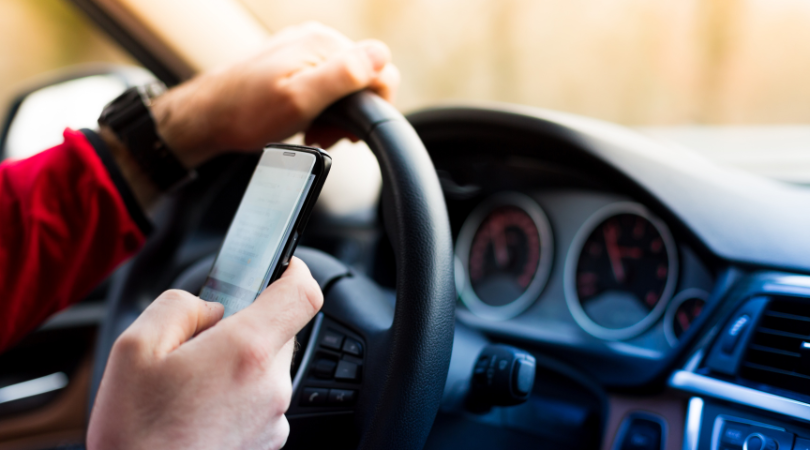 A close-up of a man's hand holding a mobile phone while the other hand is on a car steering wheel